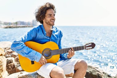 Genç İspanyol adam klasik gitar çalıyor plajda rock 'ın üzerinde oturuyor..
