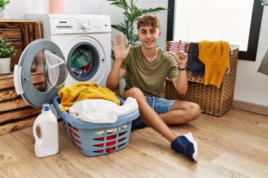 Young caucasian man putting dirty laundry into washing machine showing and pointing up with fingers number seven while smiling confident and happy. 