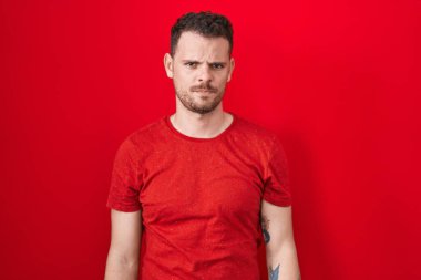 Kırmızı arka planda duran Latin asıllı genç adam şüpheci ve gergin, sorun yüzünden surat asıyor. negatif kişi. 