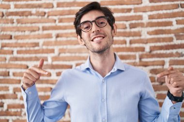 Young hispanic man standing over brick wall background looking confident with smile on face, pointing oneself with fingers proud and happy. 