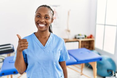 Saç örgülü siyah bir kadın ağrı kesici kliniğinde çalışıyor. Elleriyle mutlu bir jest yapıyor. Kameraya bakıp başarıyı gösteren ifade. 