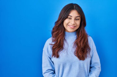 Hispanic young woman standing over blue background with hands together and crossed fingers smiling relaxed and cheerful. success and optimistic 