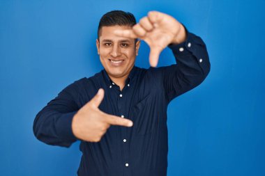 Mavi arka planda duran İspanyol genç adam gülümsüyor ve mutlu yüzlü eller ve parmaklarla çerçeve yapıyor. yaratıcılık ve fotoğrafçılık kavramı. 