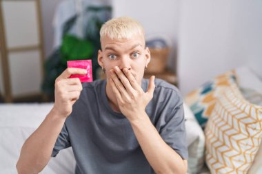 Kafkasyalı genç bir adam elinde prezervatifle yatağında oturmuş, ağzı eliyle kapatıyordu, şok olmuştu ve hata yapmaktan korkuyordu. şaşırmış ifade 