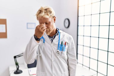 Young blond man wearing doctor uniform and stethoscope at clinic tired rubbing nose and eyes feeling fatigue and headache. stress and frustration concept. 