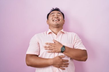 Chinese young man standing over pink background smiling and laughing hard out loud because funny crazy joke with hands on body. 