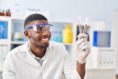 Young african american man wearing scientist uniform holding test tubes at laboratory