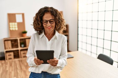 Orta yaşlı İspanyol kadın, ofiste dokunmatik ped kullanarak çalışırken kendine güvenerek gülümsüyor.