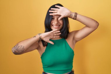 Brunette woman standing over yellow background smiling cheerful playing peek a boo with hands showing face. surprised and exited 