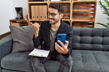 Young hispanic psychologist man doing therapy on video call with smartphone pointing thumb up to the side smiling happy with open mouth 