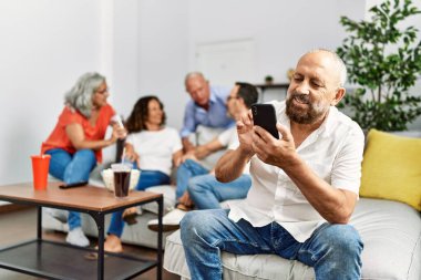 Kanepede oturan bir grup orta yaşlı arkadaş konuşuyor. Adam evde akıllı telefon kullanarak mutlu gülümsüyor..