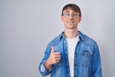 Caucasian blond man standing wearing glasses doing happy thumbs up gesture with hand. approving expression looking at the camera showing success. 