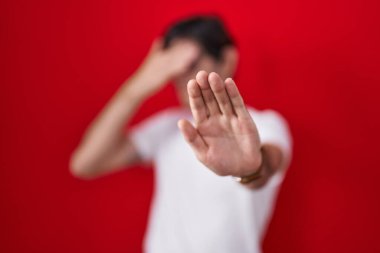 Young hispanic man standing over red background covering eyes with hands and doing stop gesture with sad and fear expression. embarrassed and negative concept. 