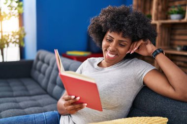 Evdeki kanepede oturan Afro-Amerikalı kadın kitap okuyor.