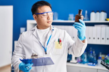 Young chinese man wearing scientist uniform reading clipboard at laboratory