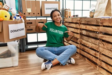 Young african american woman wearing volunteer t shirt at donations stand looking confident with smile on face, pointing oneself with fingers proud and happy. 