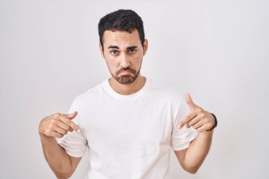 Handsome hispanic man standing over white background pointing down looking sad and upset, indicating direction with fingers, unhappy and depressed. 