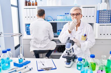 Middle age woman working at scientist laboratory covering mouth with hand, shocked and afraid for mistake. surprised expression 