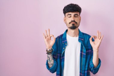 Pembe arka planda duran sakallı genç İspanyol adam rahatla ve gözün kapalı gülümse parmaklarınla meditasyon hareketi yap. yoga konsepti. 