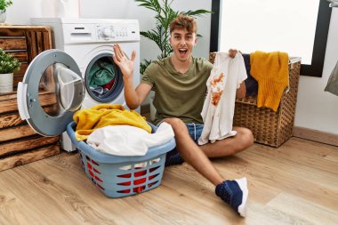 Young caucasian man doing laundry holding t shirt with stain celebrating victory with happy smile and winner expression with raised hands 