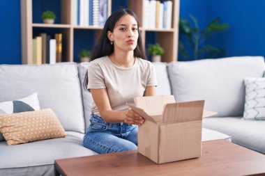 Young hispanic woman opening cardboard box looking at the camera blowing a kiss being lovely and sexy. love expression. 