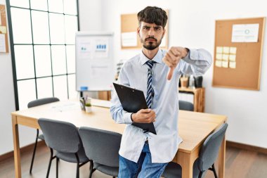 Ofiste masasında oturan sakallı, ispanyol sakallı, sinirli yüzlü, olumsuz işaretli, baş parmakları aşağıda, reddedilme konsepti olan bir adam. 