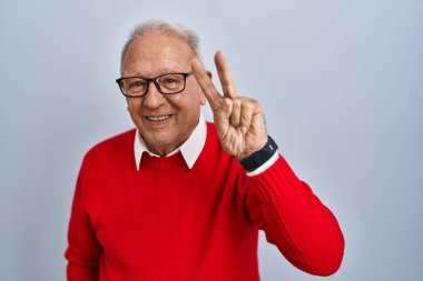 İzole edilmiş arka planda duran yaşlı adam iki numaralı parmaklarıyla gülümseyerek kendinden emin ve mutlu görünüyor.. 