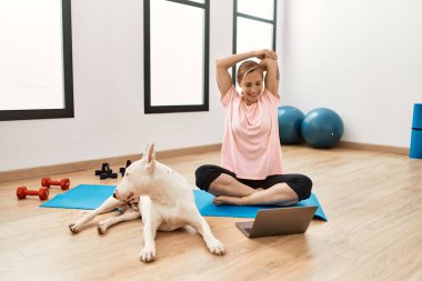 Genç beyaz kadın spor merkezinde köpekle internetten esneme dersi alıyor.