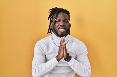 Sarı arka planda balıkçı yaka kazak giyen Afrikalı bir adam elleri bağlı yalvarıyor ve dua ediyor. Yüzünde umut ifadesi var. Çok duygusal ve endişeli. Yalvarıyor. 