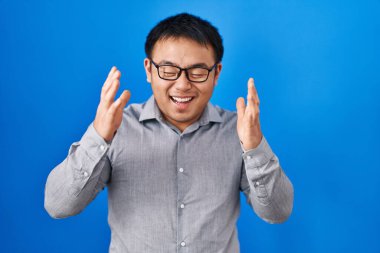 Young chinese man standing over blue background celebrating mad and crazy for success with arms raised and closed eyes screaming excited. winner concept 