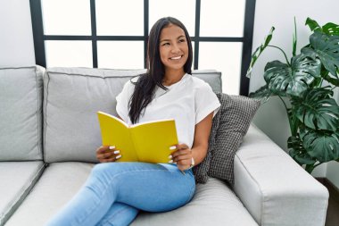 Genç Latin kadın gülümsüyor evde kendine güvenen kitap okuyor.