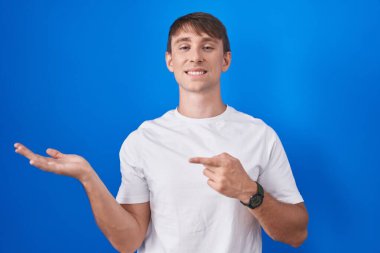 Caucasian blond man standing over blue background amazed and smiling to the camera while presenting with hand and pointing with finger. 