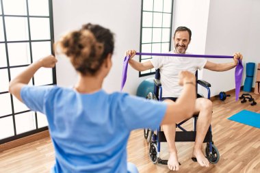 Orta yaşlı bir adam ve kadın fizik tedavi kliniğinde tekerlekli sandalyede oturan elastik bant kullanarak rehabilitasyon seansı yapıyorlar.