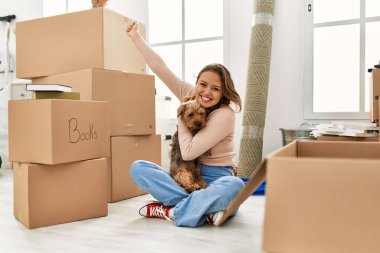 Genç, güzel, İspanyol bir kadın köpeğini kucaklıyor. Yeni evinde neşeli bir ifadeyle yerde oturuyor.