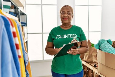 Senior african american woman wearing volunteer uniform writing on clipboard at charity center