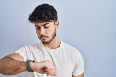 Sakallı İspanyol bir adam beyaz arka planda duruyor. Saati kontrol ediyor, rahat ve kendinden emin. 