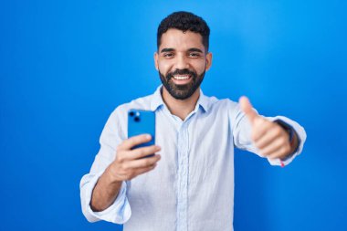 Hispanic man with beard using smartphone typing message approving doing positive gesture with hand, thumbs up smiling and happy for success. winner gesture. 
