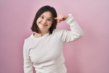 Down sendromlu bir kadın pembe arka planda durup gülümseyerek tek parmağıyla kafasını işaret ediyor. Harika bir fikir ya da düşünce, iyi bir hafıza. 