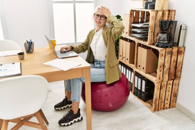 Ofiste çalışan beyaz, güzel bir iş kadını pilates topunda oturuyor. Elleri havada, şaşırmış ve sinirli bir halde. 