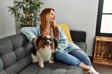Beyaz bir kadın, evinde köpekle kanepede otururken kendinden emin bir şekilde gülümsüyor.