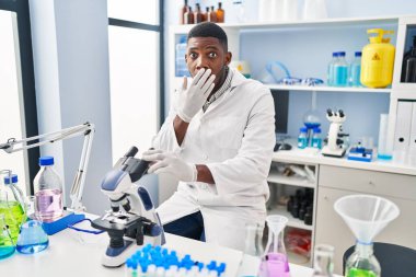 African american man working at scientist laboratory covering mouth with hand, shocked and afraid for mistake. surprised expression 