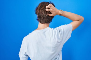 Latin kökenli genç adam mavi arka planda geriye doğru duruyor. Elleri kafasında şüpheyi düşünüyor. 