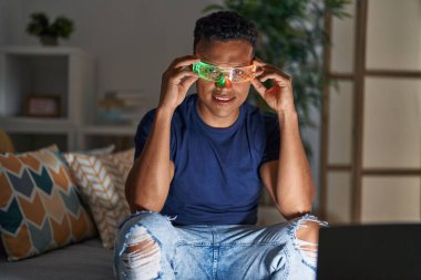 Young latin man using virtual reality glasses sitting on sofa at home