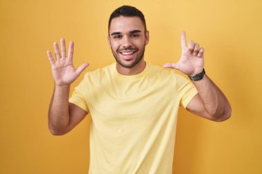 Sarı arka planda duran İspanyol genç adam yedinci parmaklarını gösterip işaret ederken kendinden emin ve mutlu bir şekilde gülümsüyor.. 