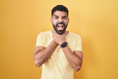 Hispanic man with beard standing over yellow background shouting suffocate because painful strangle. health problem. asphyxiate and suicide concept. 