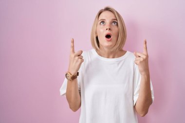 Young caucasian woman standing over pink background amazed and surprised looking up and pointing with fingers and raised arms. 