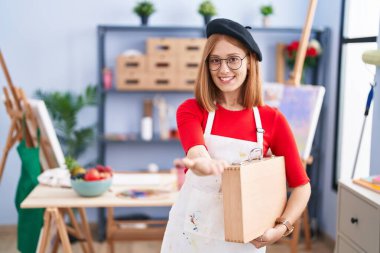 Young redhead woman at art studio holding art case smiling cheerful offering palm hand giving assistance and acceptance. 