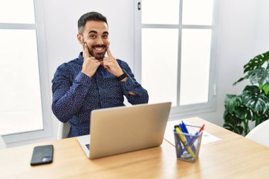 Ofiste sakallı, laptopuyla çalışan ağzı açık gülümseyen, parmakları işaret eden ve neşeli gülümsemeye zorlayan genç İspanyol bir adam. 