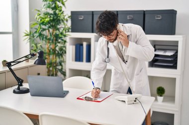 Young hispanic man wearing doctor uniform talking on telephone writing on document at clinic