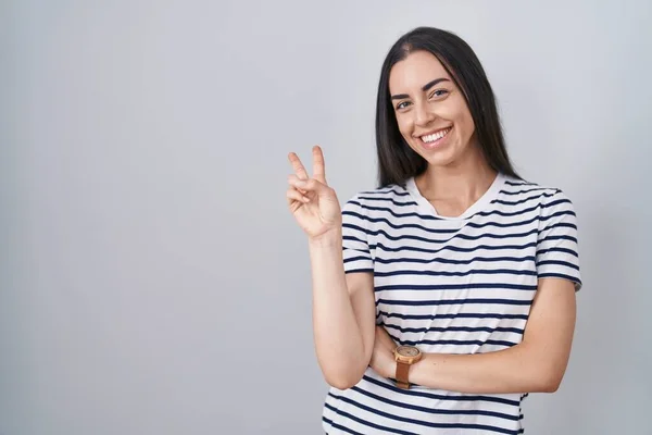 Young brunette woman wearing striped t shirt smiling with happy face winking at the camera doing victory sign with fingers. number two.